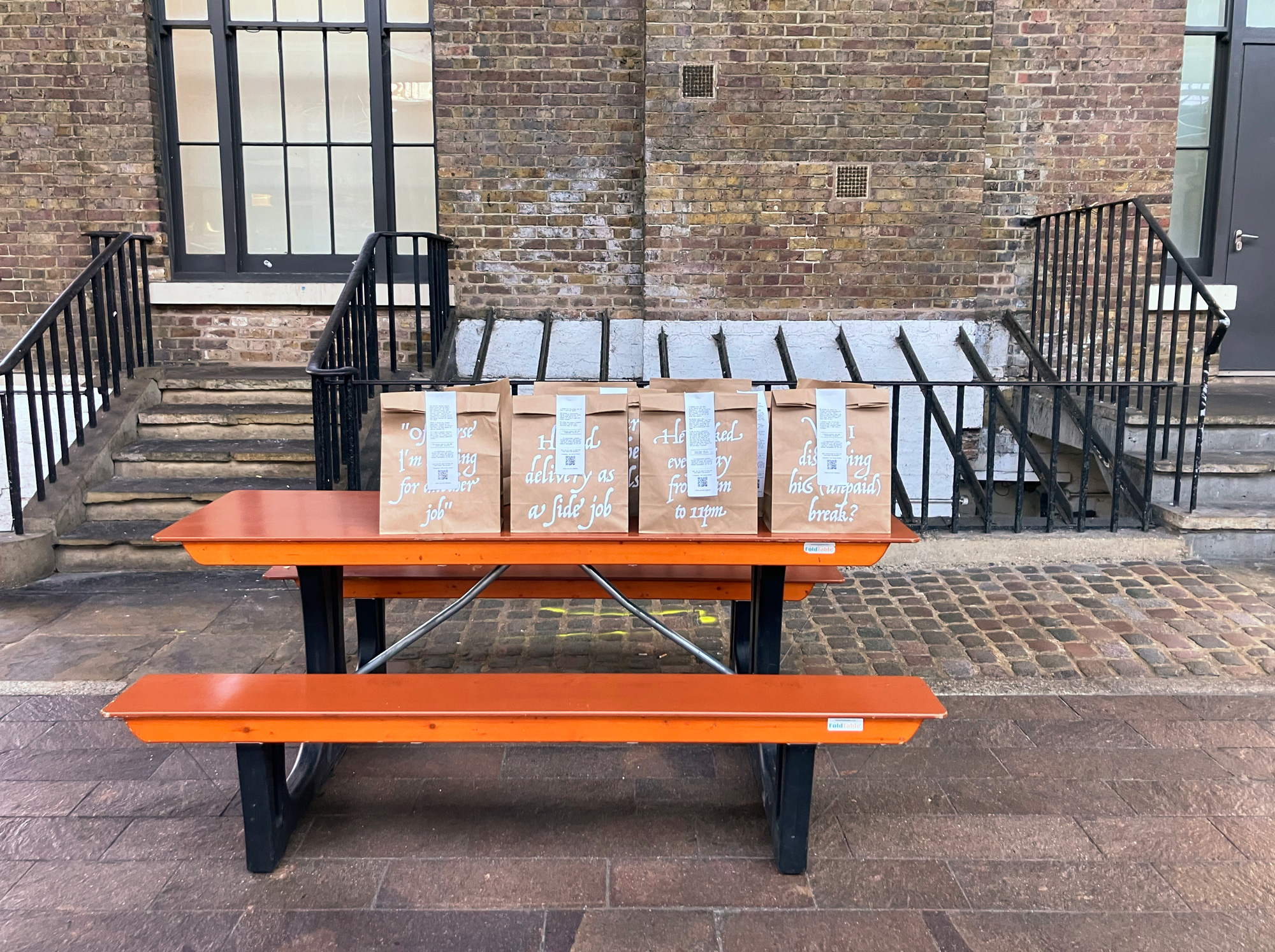 Ten kraft paper bags with phrases printed on them and receipts stapled to them on top of a canteen table.